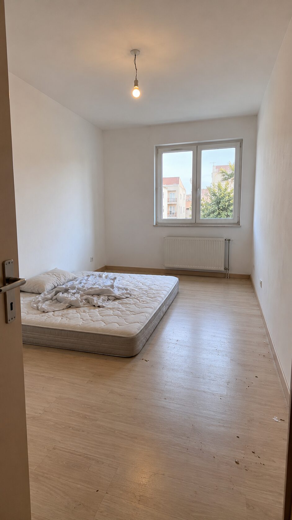 Bare rental bedroom with mattress on the floor, white walls, beige laminate and a bare ceiling bulb